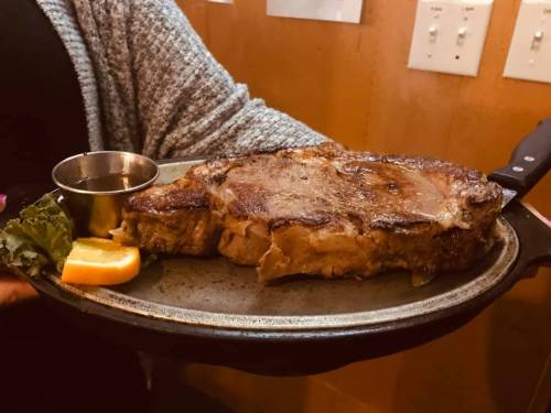 A person holds a large, juicy steak on a platter, garnished with lettuce and an orange slice, with a small metal cup nearby.
