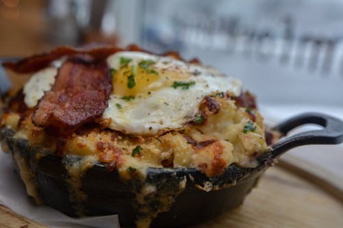 A skillet dish featuring creamy mac and cheese topped with crispy bacon and a sunny-side-up egg, garnished with parsley.