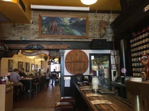 Interior of a rustic restaurant with wooden decor, a large painting, and a bar area. People are dining in the background.