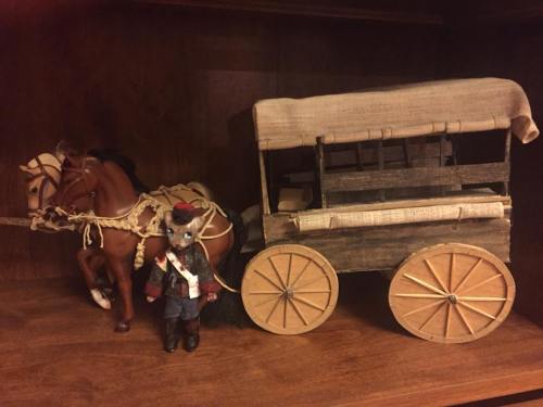 A wooden toy wagon with wheels, pulled by a horse, alongside a small doll dressed in a black coat and hat.