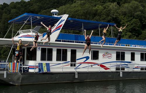 A group of six people in life jackets jumping off a houseboat into the water, surrounded by green trees.