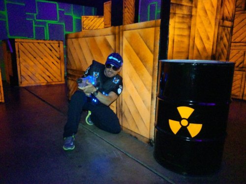 A person in a black vest poses next to a yellow and black radioactive barrel in a colorful, illuminated environment.
