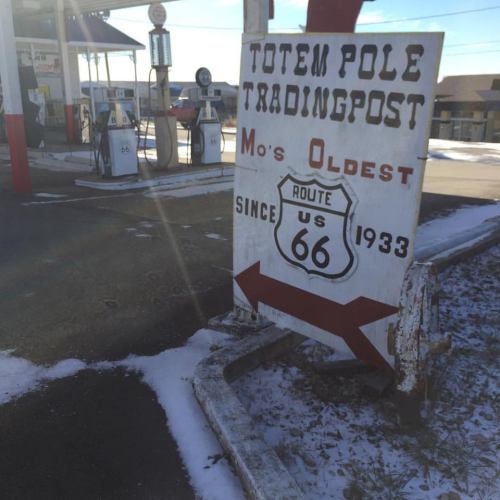 Sign for Totem Pole Trading Post, featuring Route 66, with vintage gas pumps in a snowy setting.