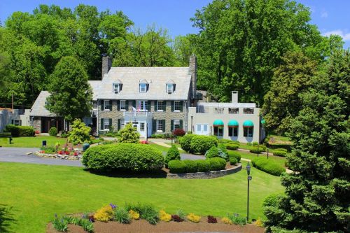 A large, elegant house surrounded by lush greenery and colorful flower beds on a sunny day.