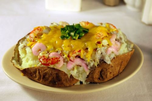 A baked potato topped with shrimp, melted cheese, and green onions on a white plate.