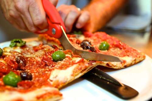 A person using scissors to cut a pizza topped with olives and green peppers on a white plate.