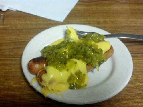A hot dog on a plate topped with yellow mustard and chopped pickles, served with a fork on a wooden table.