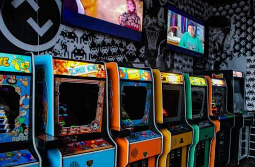 A row of colorful vintage arcade machines in a retro-themed gaming room with screens displaying videos.