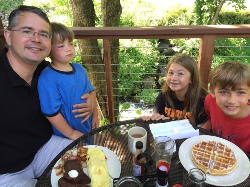 A family enjoys breakfast on a patio, featuring waffles, an omelet, and a scenic view of greenery.