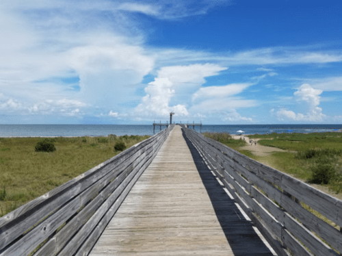 A wooden pier extends into the ocean under a bright blue sky with fluffy clouds, surrounded by green grass.