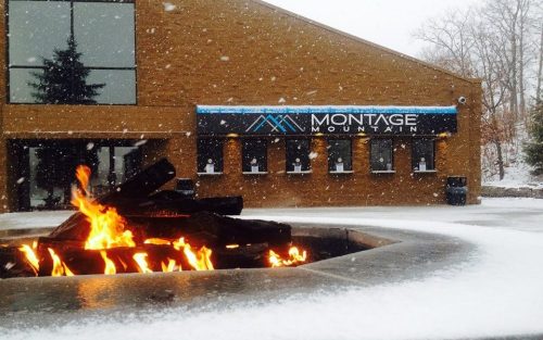 A snowy scene at Montage Mountain, featuring a fire pit in the foreground and a building with a sign in the background.