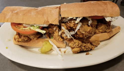 A large sandwich filled with fried chicken, lettuce, tomato, and pickles, served on a toasted hoagie roll.
