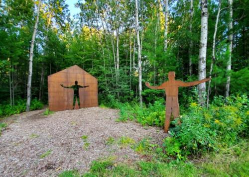 A metal silhouette stands with arms outstretched beside a rusted house structure in a wooded area.