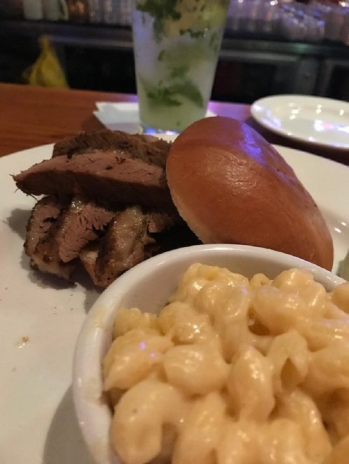 A plate featuring sliced grilled meat, a bun, and a side of creamy macaroni and cheese, with a drink in the background.