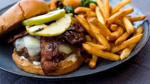 A juicy burger topped with grilled apple, bacon, and cheese, served with crispy fries and a side of mixed greens.