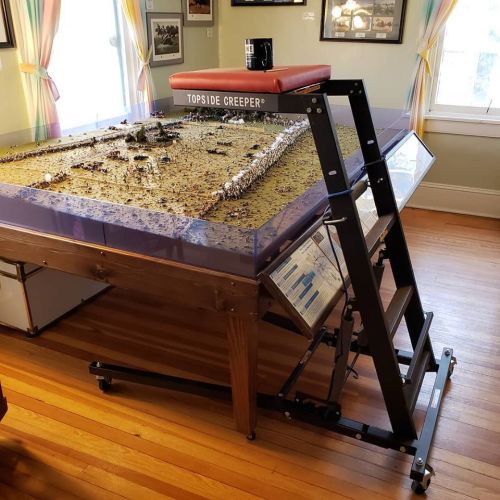 A model display table with a clear cover, featuring a detailed landscape, and a ladder beside it in a well-lit room.