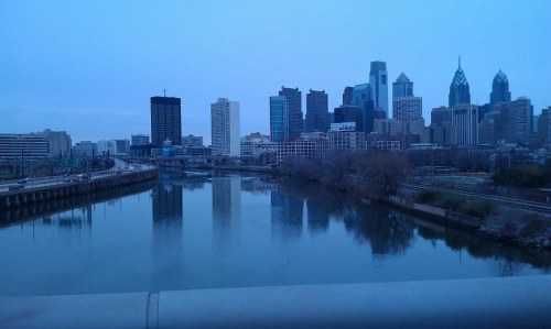City skyline of Philadelphia at dusk, reflecting in the calm river with bare trees along the shore.