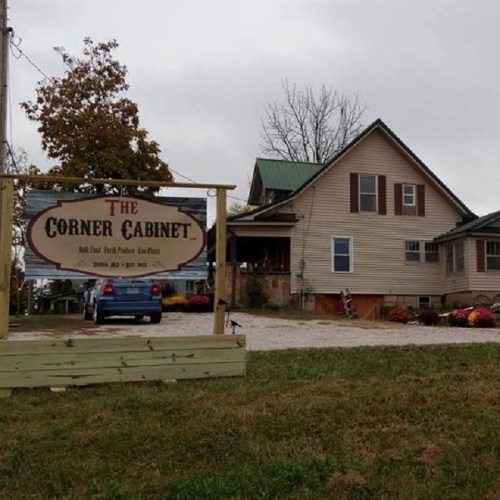 Sign for "The Corner Cabinet" in front of a house with a green roof, surrounded by trees and flowers.