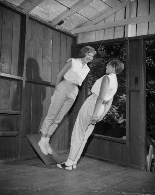 Two women in vintage clothing pose playfully in a wooden cabin, one leaning against the wall and the other standing on a slanted board.