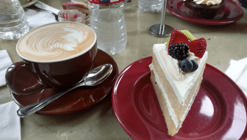 A cup of latte art coffee beside a slice of layered cake topped with berries on a red plate.