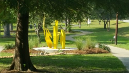 A bright yellow abstract sculpture stands in a park, surrounded by greenery and a winding path.