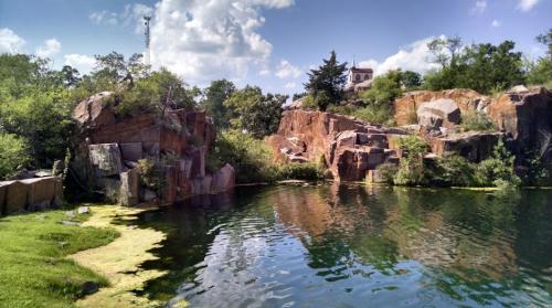 A serene quarry pond surrounded by rocky cliffs and lush greenery under a blue sky with fluffy clouds.