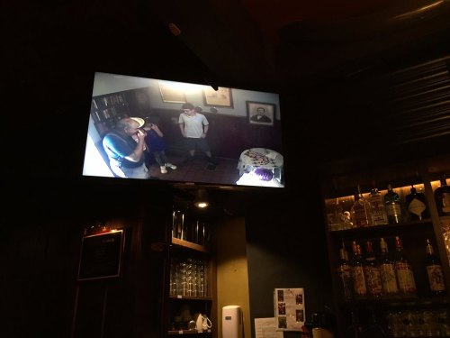 A TV screen displays a live feed of two people standing in a dimly lit room with bookshelves and a portrait on the wall.