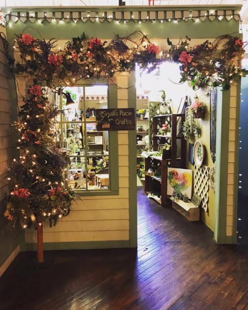 A charming craft shop entrance adorned with flowers and lights, leading to a cozy, decorated interior.