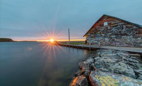 A colorful, graffiti-covered building by a calm waterway at sunset, with a starburst effect from the sun.
