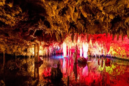 A colorful cave interior with stalactites and stalagmites reflected in still water, illuminated by vibrant lights.