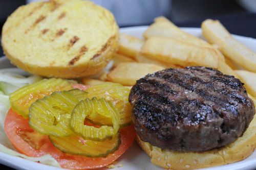 A grilled hamburger patty with lettuce, tomato, pickles, and fries on a plate, with a toasted bun on the side.