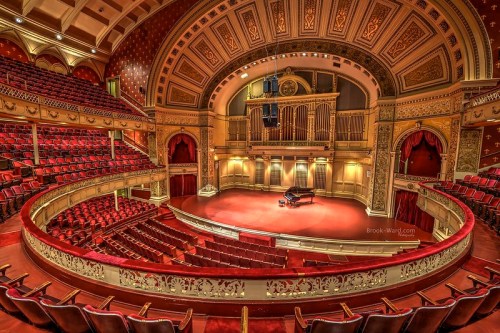 A grand concert hall with red velvet seats, ornate architecture, and a grand piano on stage, illuminated and ready for performance.