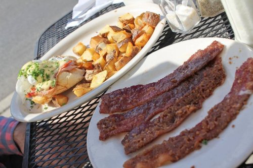 A plate of crispy bacon strips next to a dish of seasoned potatoes and a sandwich topped with sauce and herbs.