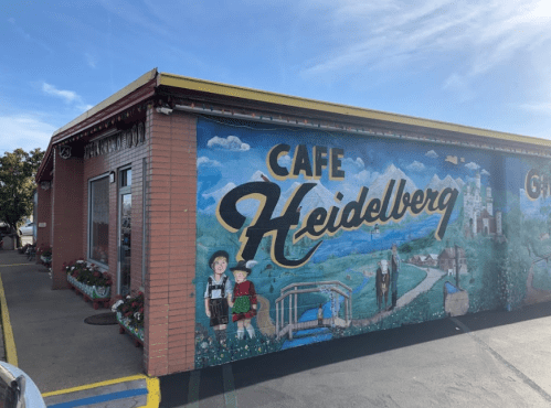 Mural on the side of Cafe Heidelberg featuring scenic landscapes and traditional figures. Bright blue sky above.