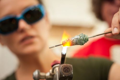 A person wearing sunglasses shapes molten glass with a tool, surrounded by flames in a glassblowing workshop.