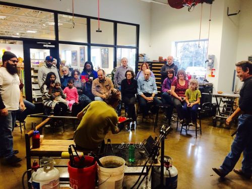 A diverse group of people watches a glassblowing demonstration in a workshop setting.