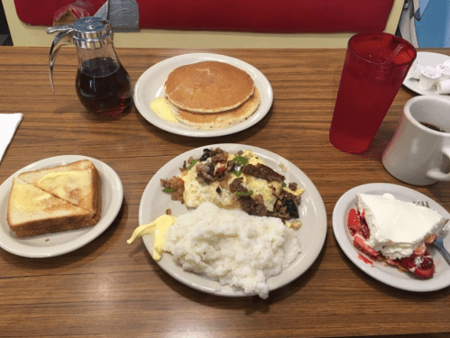 A breakfast spread featuring pancakes, toast, grits, an omelet, and a dessert with strawberries and whipped cream.