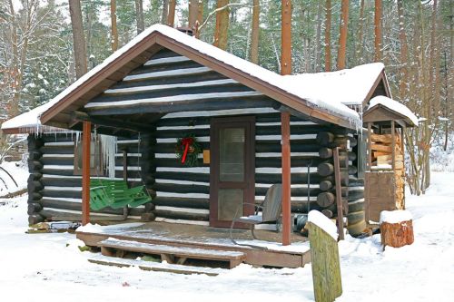 A cozy log cabin in a snowy forest, featuring a porch with a green swing and a festive wreath on the door.