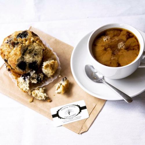 A muffin with blueberries, partially eaten, next to a cup of coffee with cream swirling, on a white tablecloth.
