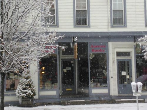 A snowy scene of a quaint bookstore with a sign reading "Aarons Books" and a tree in front.
