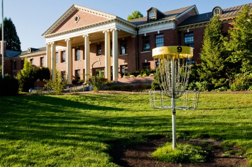 A disc golf basket in front of a large brick building with columns and landscaped greenery.
