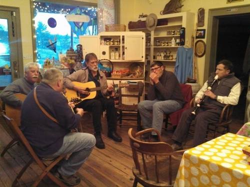 Five musicians play instruments in a cozy room filled with shelves of jars and decorations, creating a lively atmosphere.