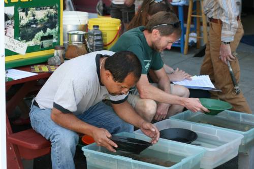 Two men pan for gold in shallow containers at an outdoor event, with informational materials and tools nearby.