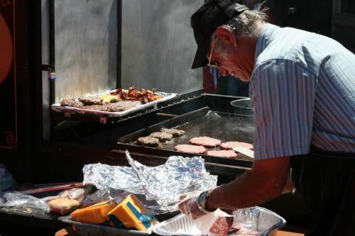 A man cooks burgers and sausages on a grill, preparing food at an outdoor event. Smoke rises from the grill.