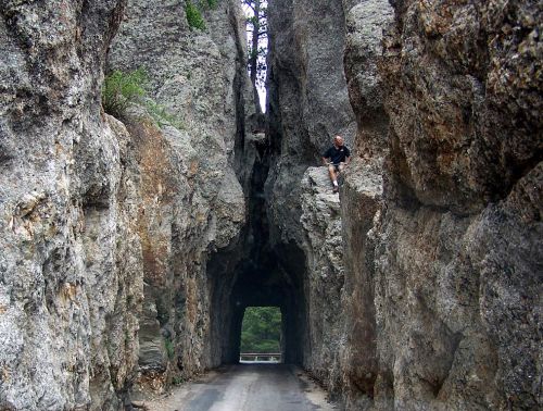 A narrow road cuts through towering rock formations, with a person perched on a ledge above.