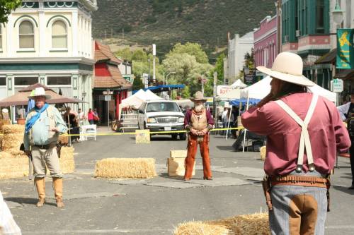Three cowboys in Western attire stand in a street lined with hay bales, with tents and a truck in the background.