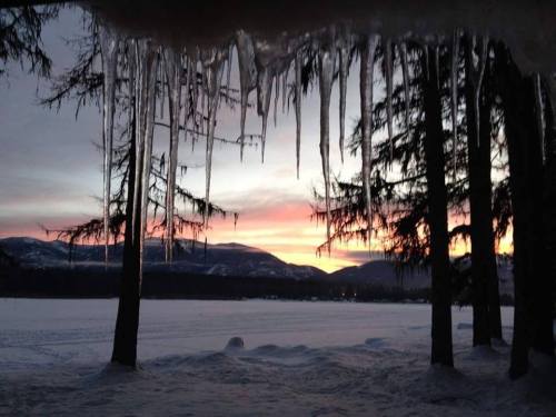 Icicles hang from trees at sunset, with snow-covered mountains and a frozen lake in the background.