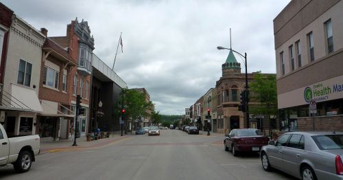 A view of a small town street with shops, cars, and cloudy skies, showcasing a mix of historic and modern architecture.