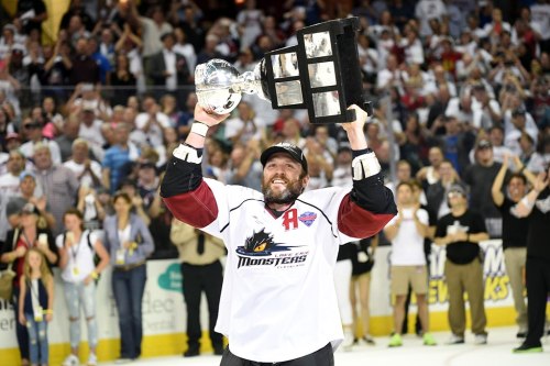 A hockey player in a white jersey lifts a championship trophy, celebrating in front of a cheering crowd.