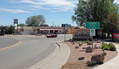 A road sign welcomes visitors to Alamosa, with a restaurant and trees in the background on a sunny day.